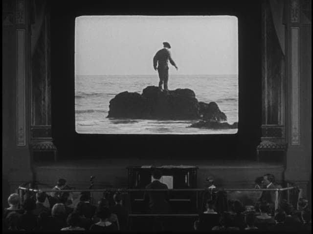 A cinema audience watches Buster Keaton stranded on a rock in the sea, on screen above the pianist, in Sherlock Jr. (1924)
