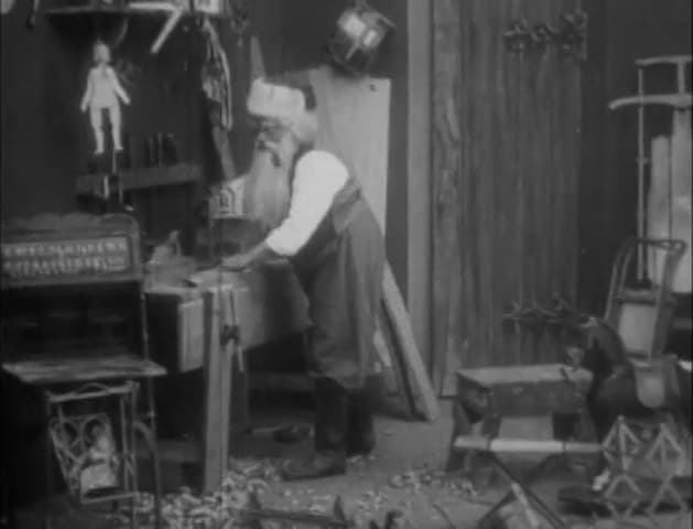 Santa Claus works at a bench in a toy workshop hung with dolls and sleds in The Night Before Christmas (1905)
